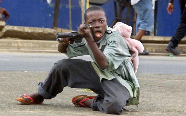 A child soldier wearing a teddy bear backpack points his gun at the photographer in a street of Monrovia, Liberia / AP